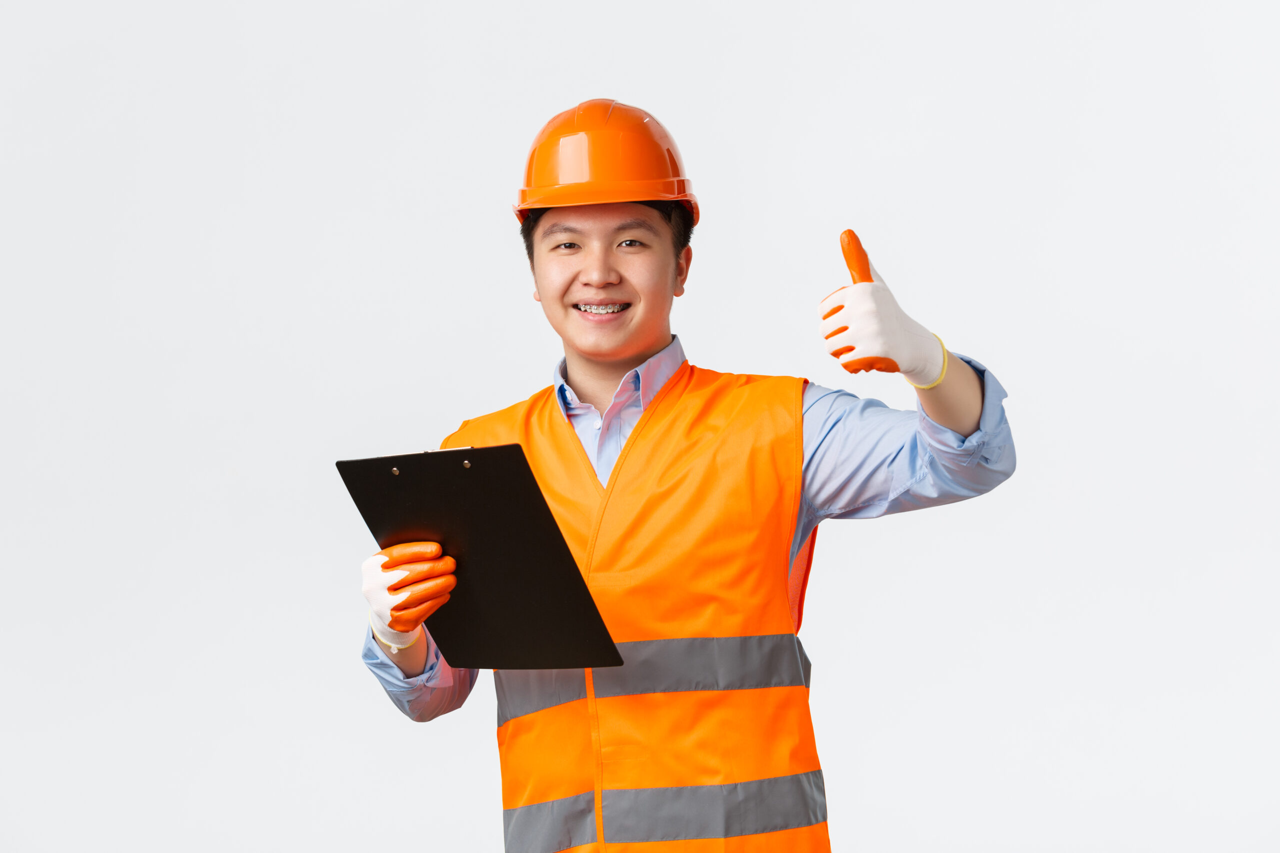 Building sector and industrial workers concept. Smiling satisfied asian chief engineer, architect making inspection showing thumbs-up in approval, holding clipboard with documents, wear helmet.